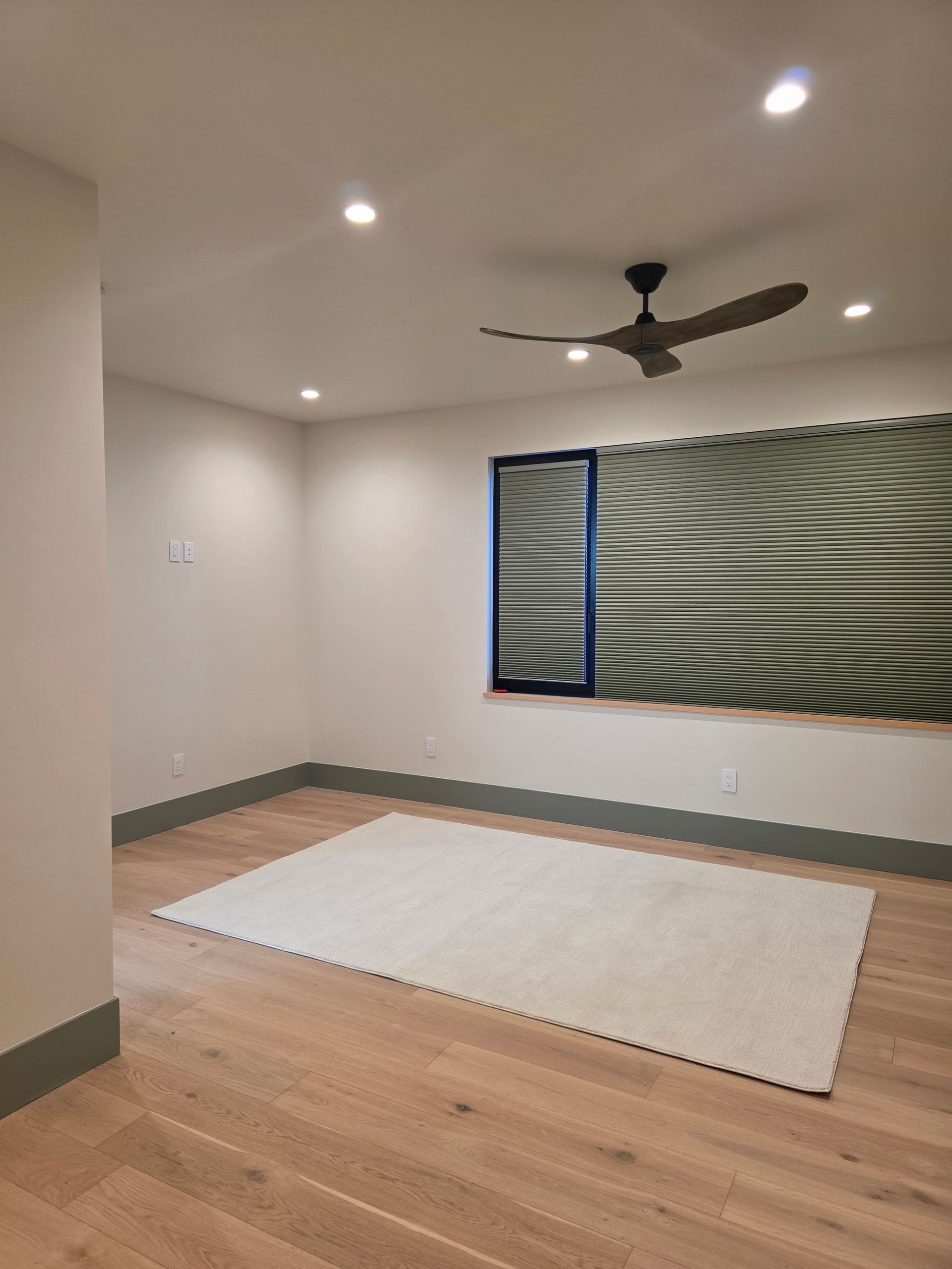 Empty room with wood floor, white rug, ceiling fan, recessed lights, and a window with blinds.