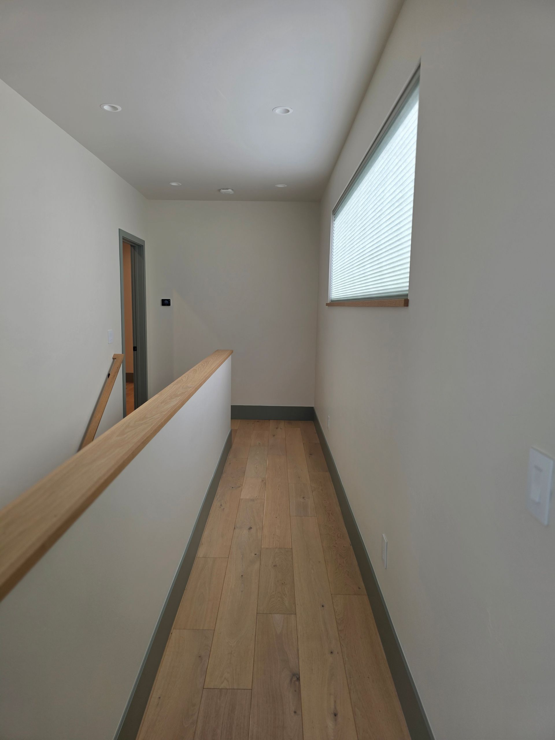 Narrow upstairs hallway with light wood floor, white walls, window blind, and wooden railing.