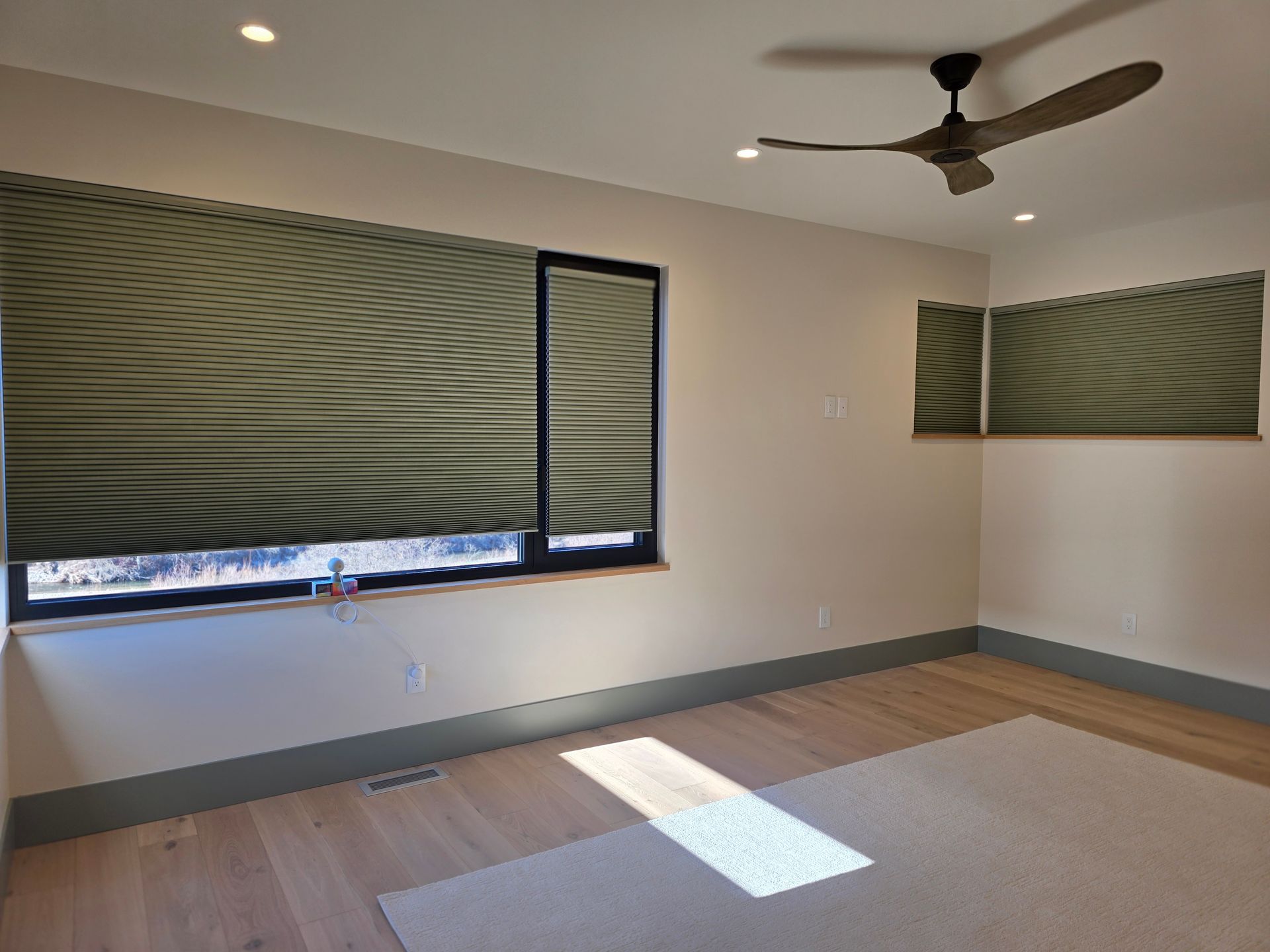 Empty room with beige walls, wood floor, ceiling fan, and large windows with green blinds.