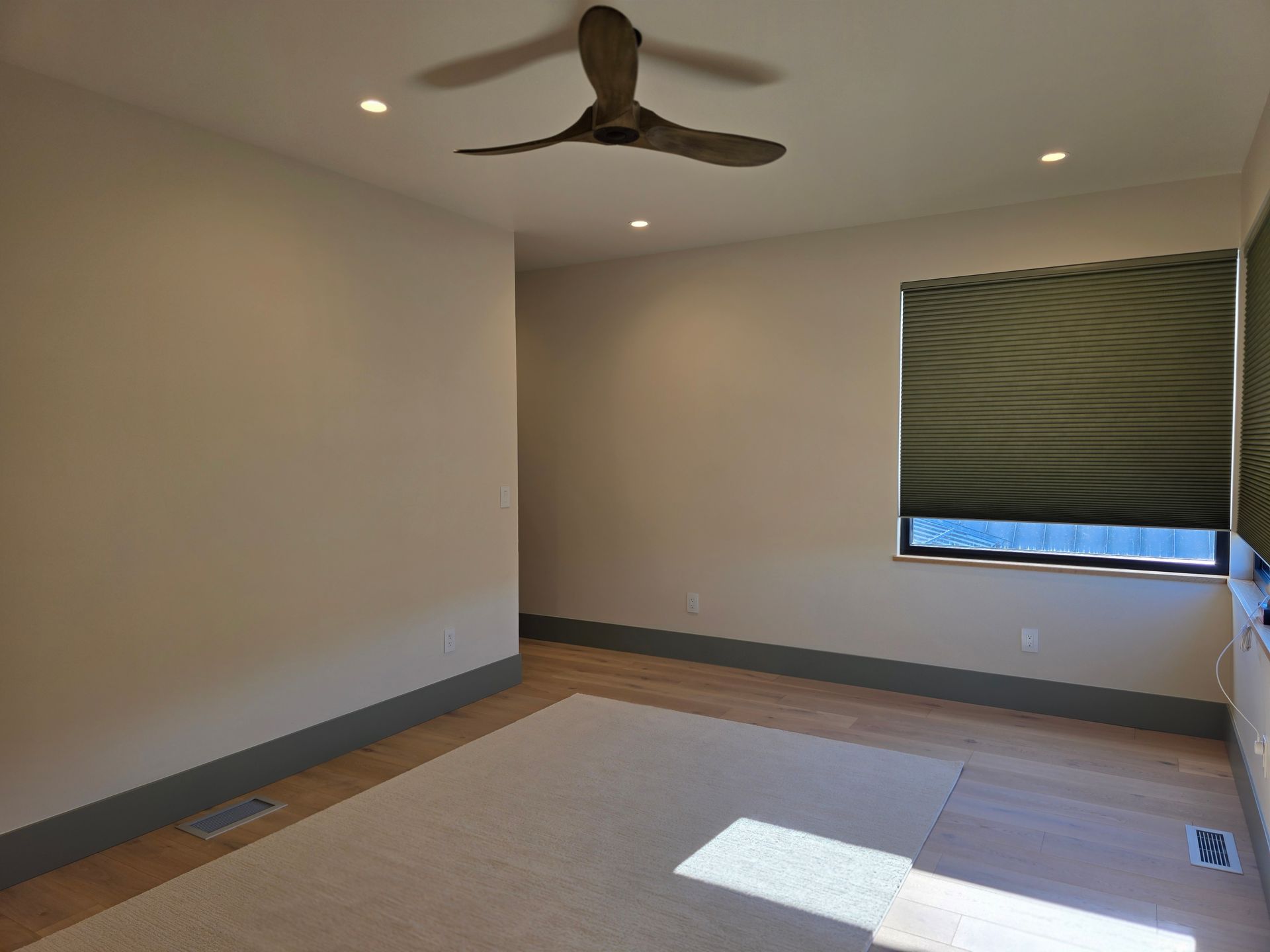 Empty beige bedroom with ceiling fan, window blinds, and sunlight on carpeted floor
