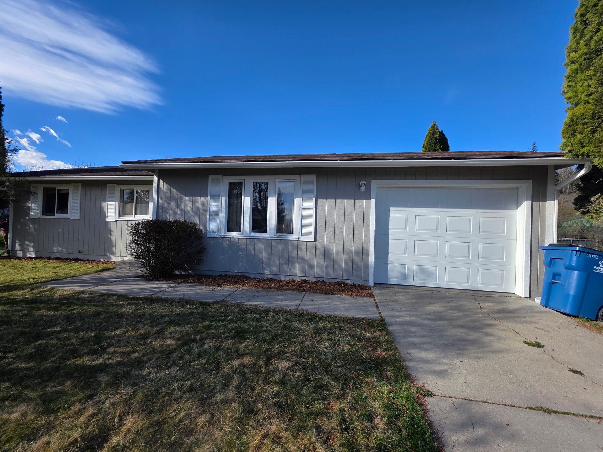 Single-story gray house with white trim, attached garage, and blue recycling bin on a sunny day
