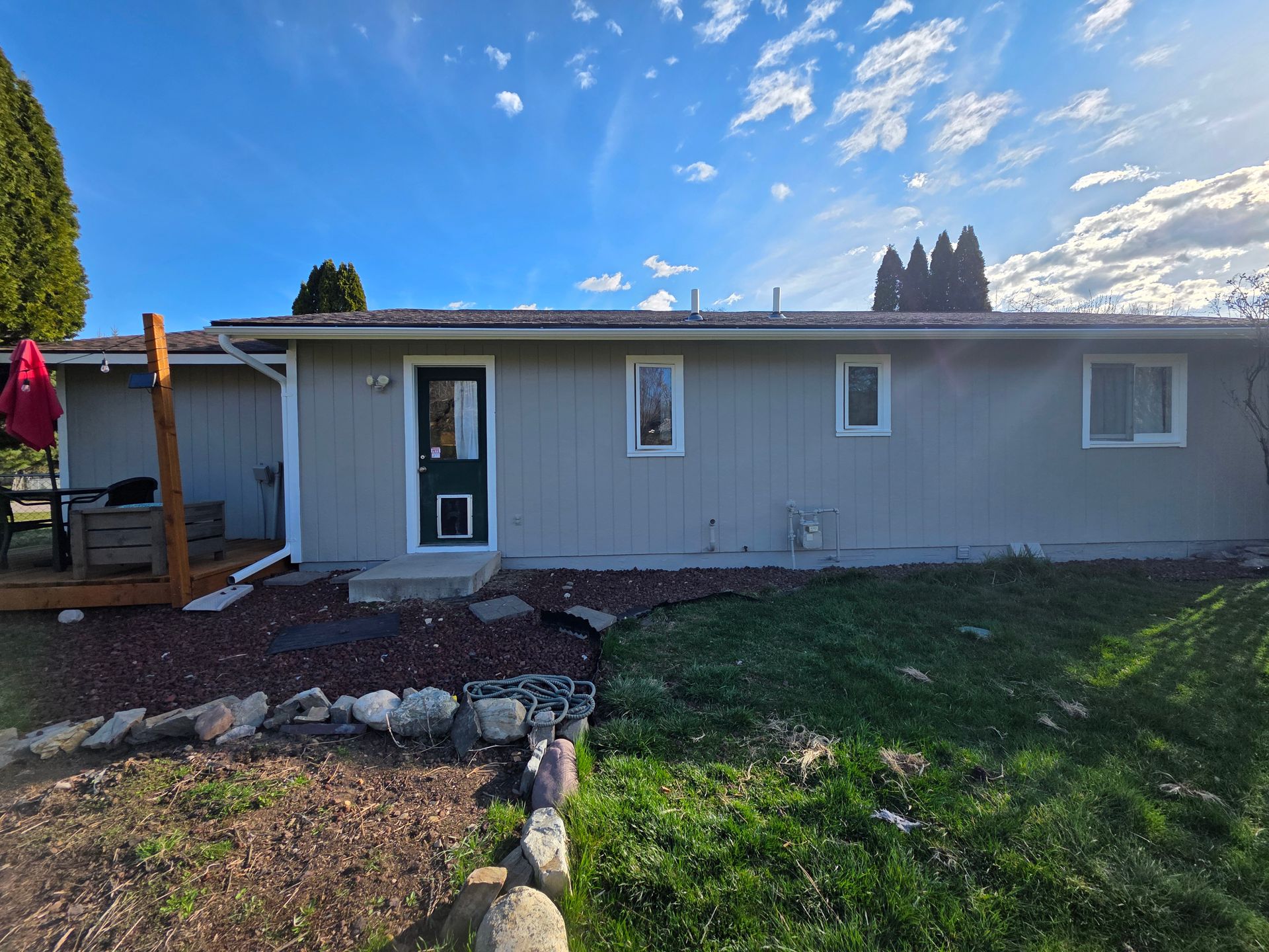Single-story gray house with a white door, front yard, and rock border under a blue sky