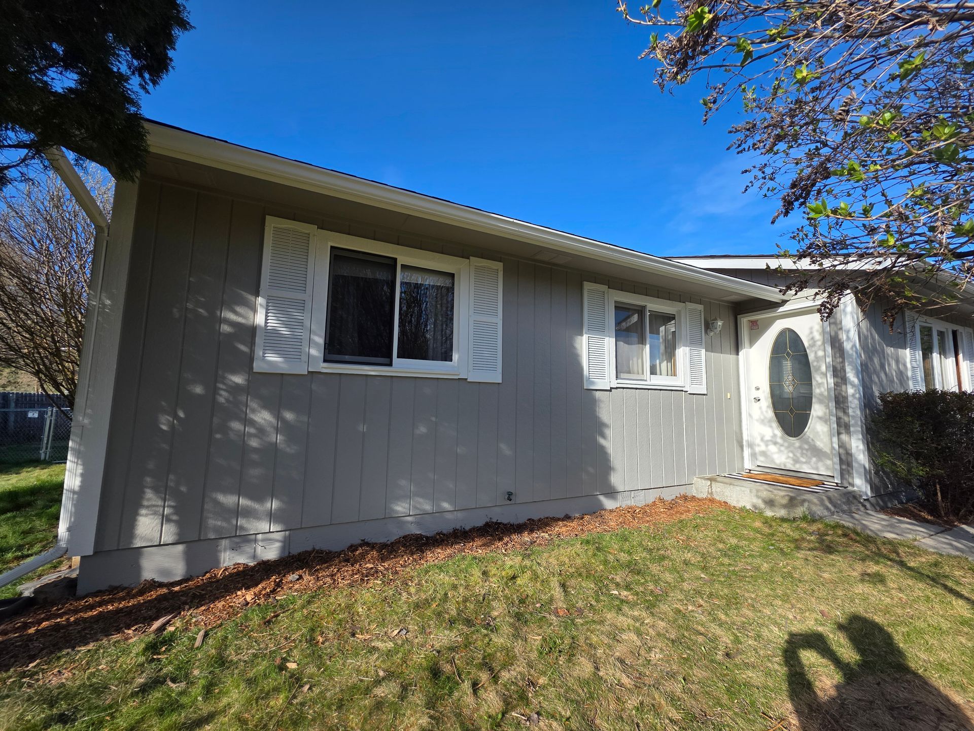 Gray house with white shutters and oval front door on a sunny lawn under a blue sky