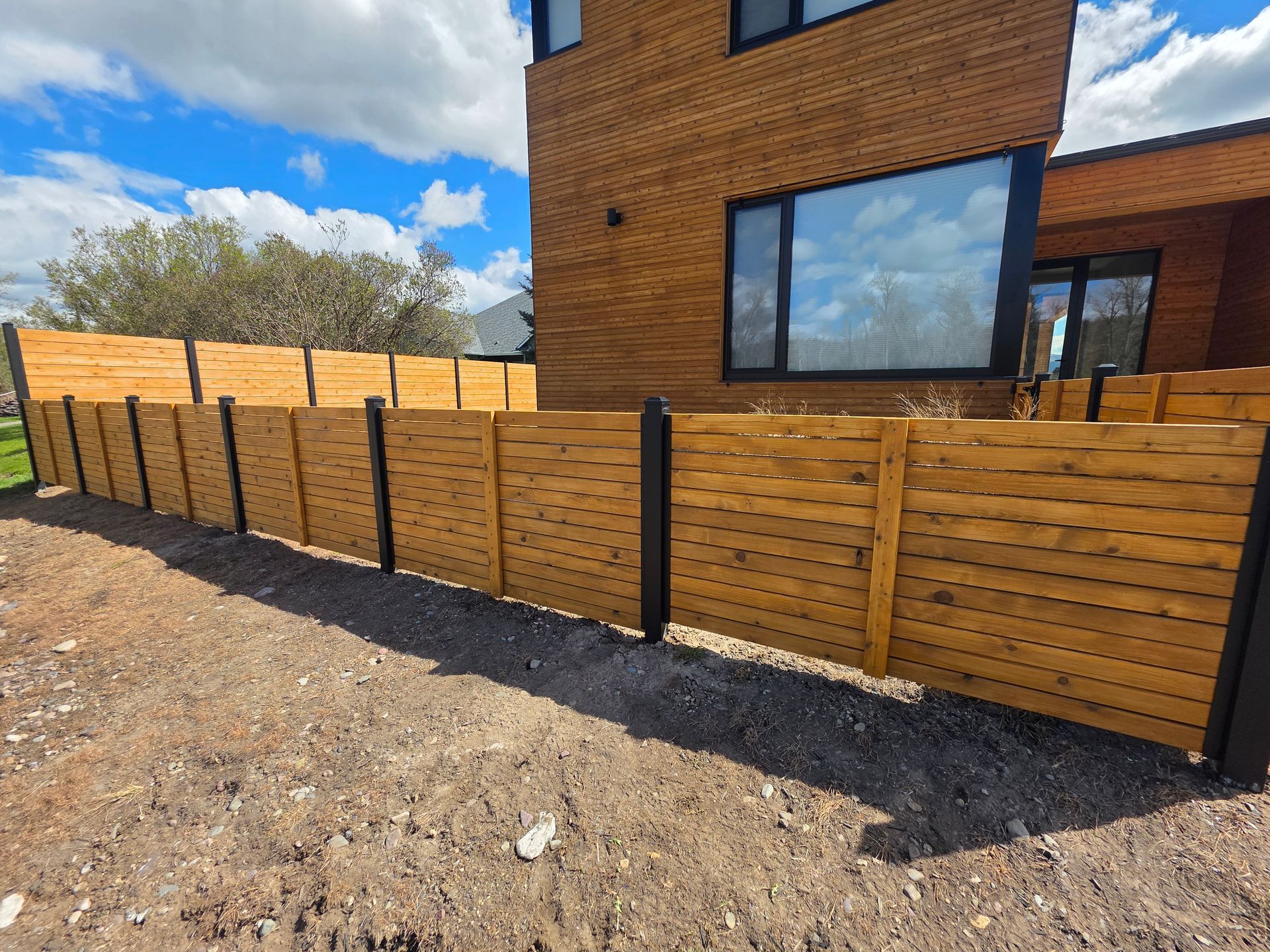 Wooden slat fence along a gravel yard beside a modern brick house under a blue sky