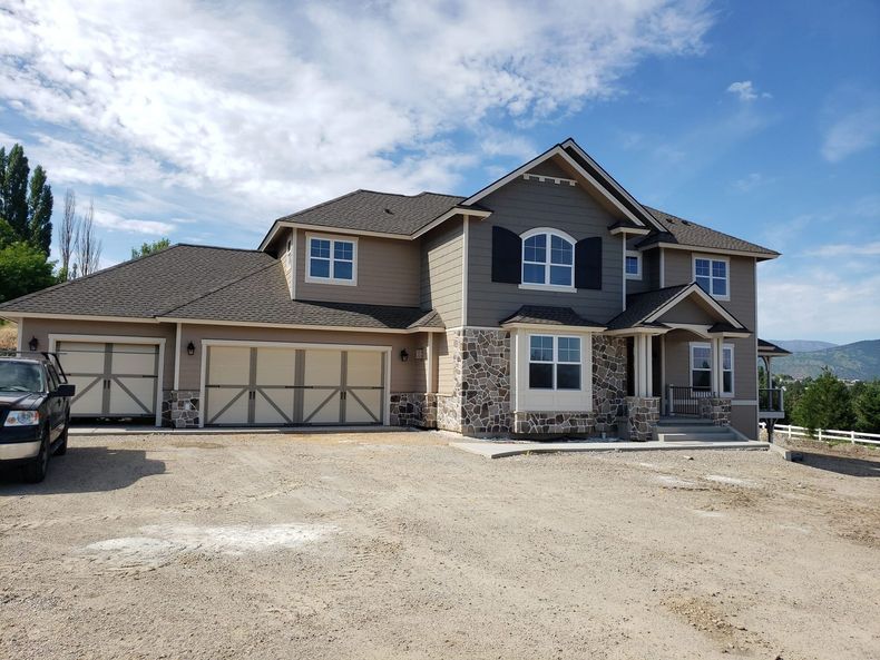 Large beige suburban house with three-car garage, stone accents, and gravel driveway under a blue sky