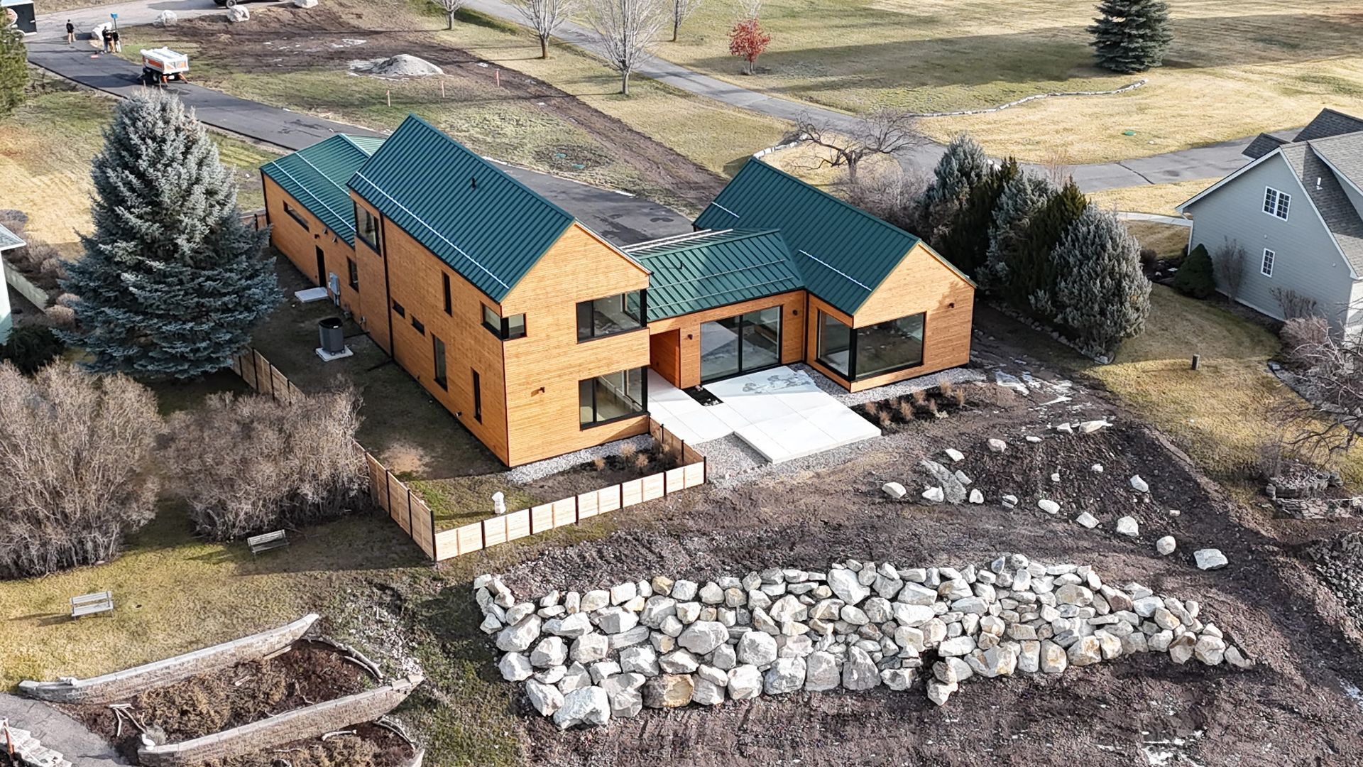Aerial view of a tan house with green roof beside rocks, trees, and a grassy yard.