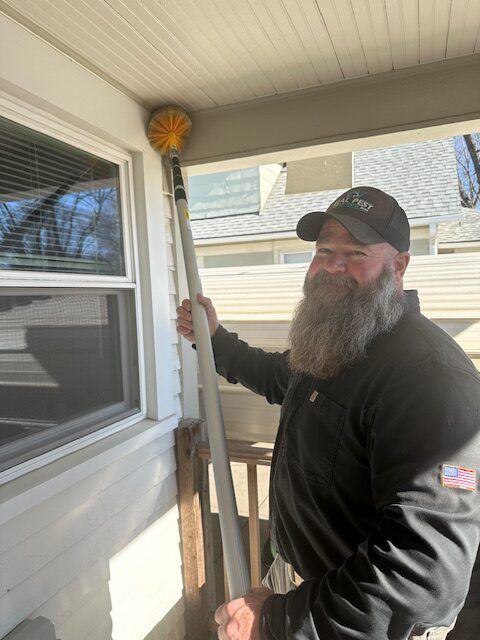 A bearded person smiles while using a long-handled brush to clean a cobweb from a porch ceiling corner.