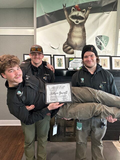 Three staff members in company uniforms hold a framed certificate, with one person lifted horizontally in the air.