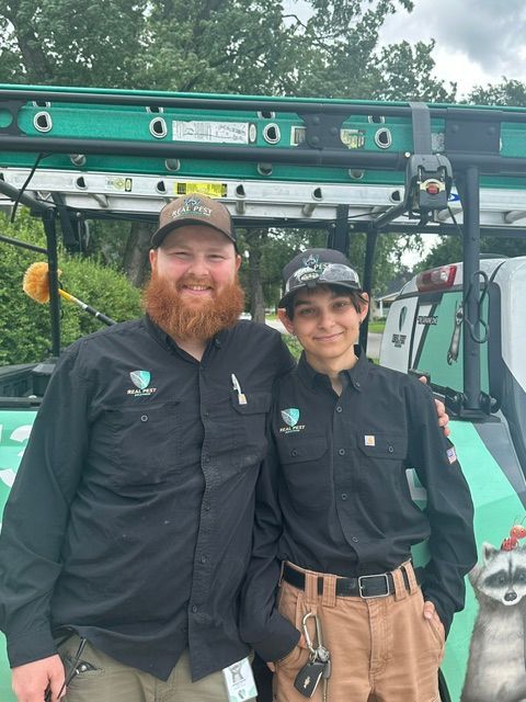 Two staff members in black uniforms stand beside a green utility truck, smiling outdoors.