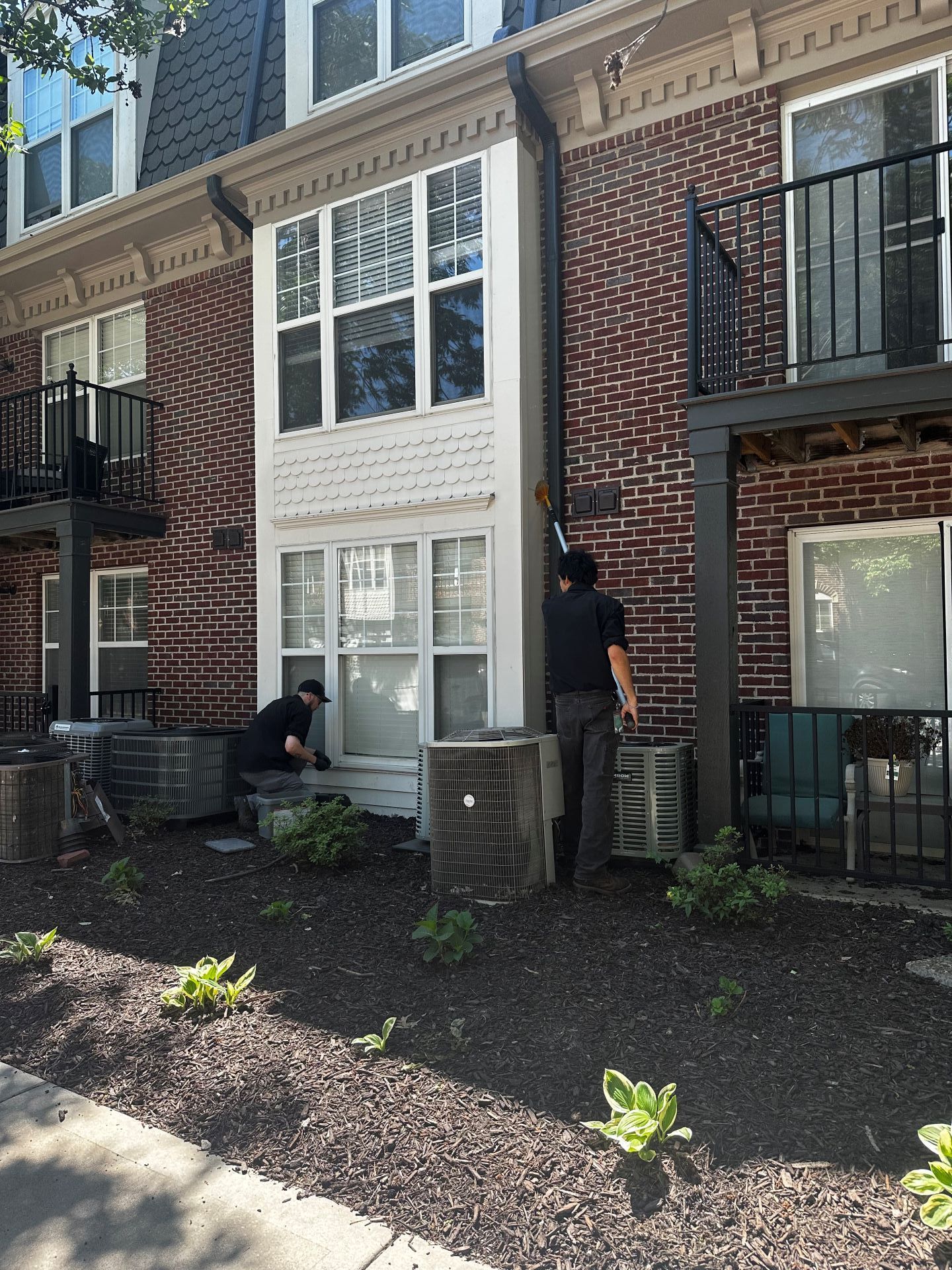 Brick townhouse patio with two people near a white-trimmed door and balcony, surrounded by mulch and potted plants