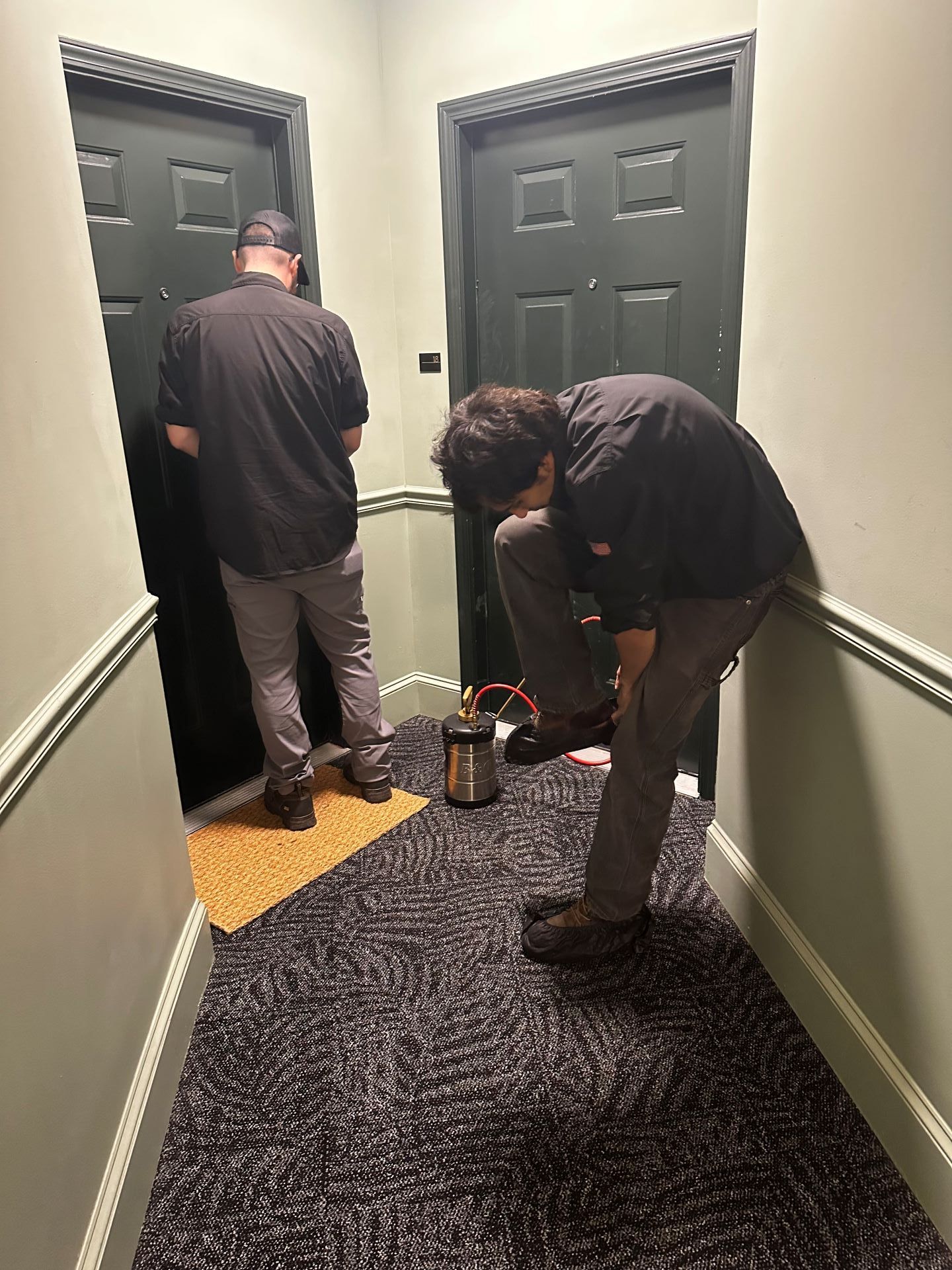 Two people standing in a hallway, one working near the floor by a doorway.