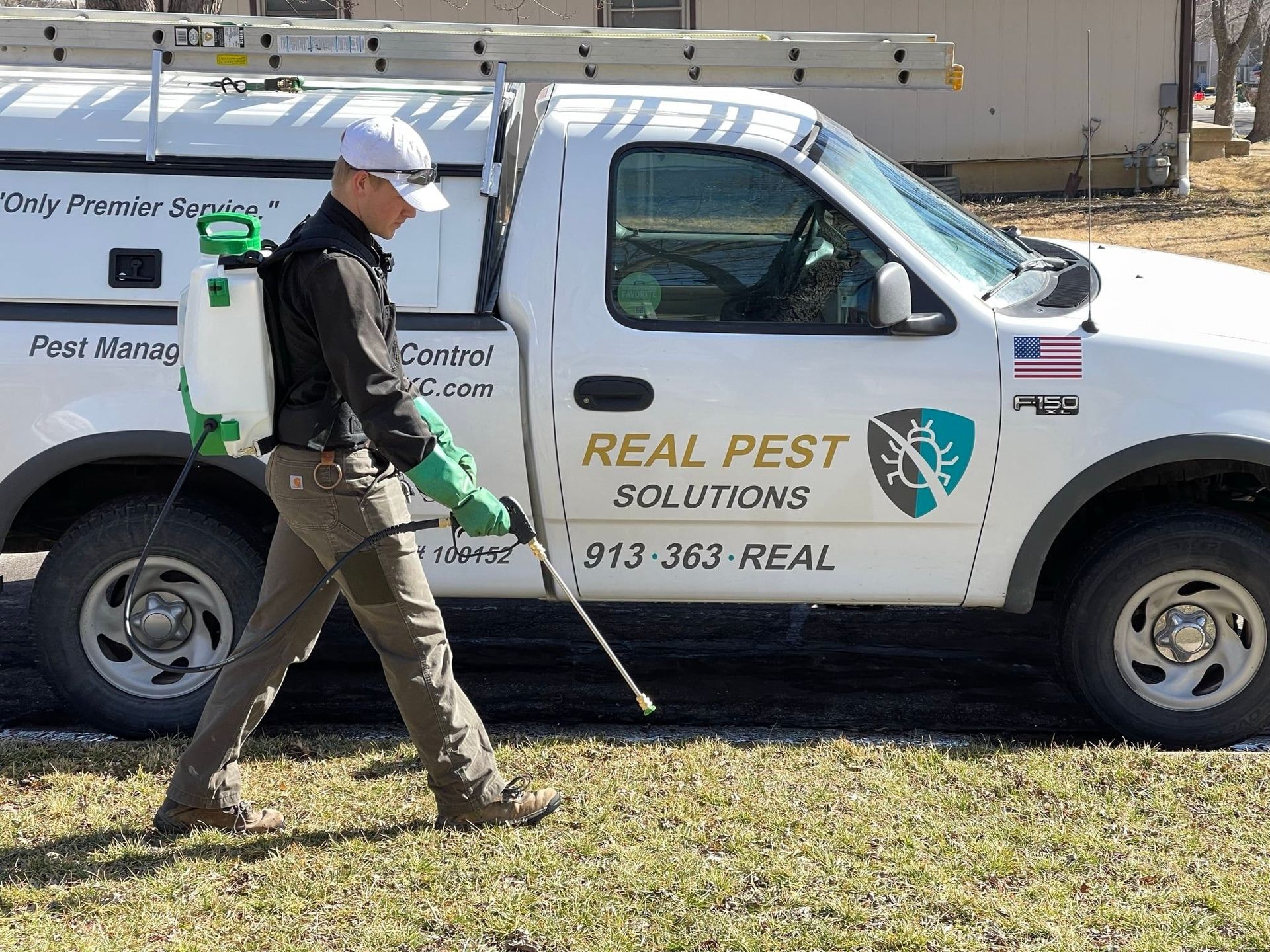 A technician with a backpack sprayer walks beside a Real Pest Solutions truck parked on a sunny, grassy lawn.