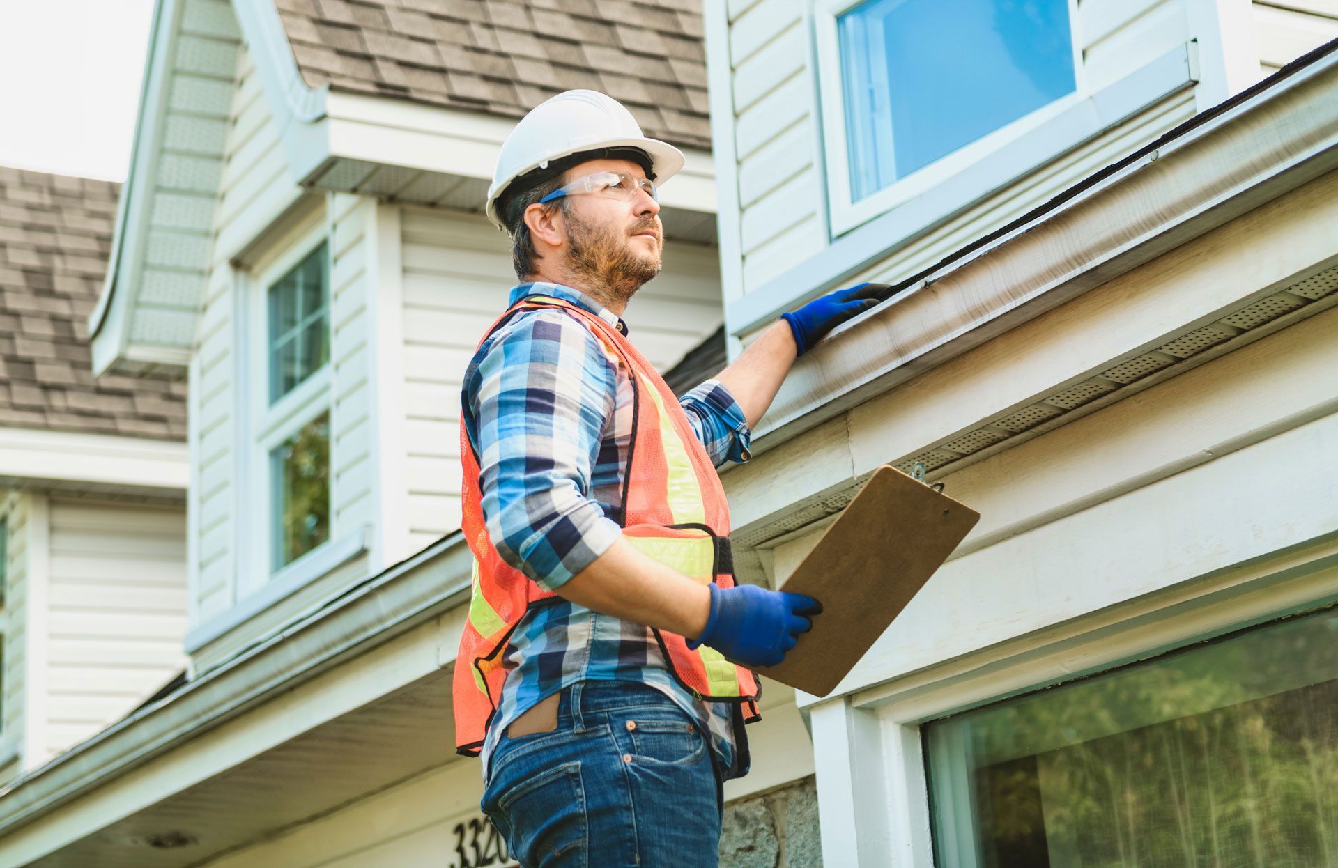 Worker in hard hat inspecting house siding with clipboard and tool belt