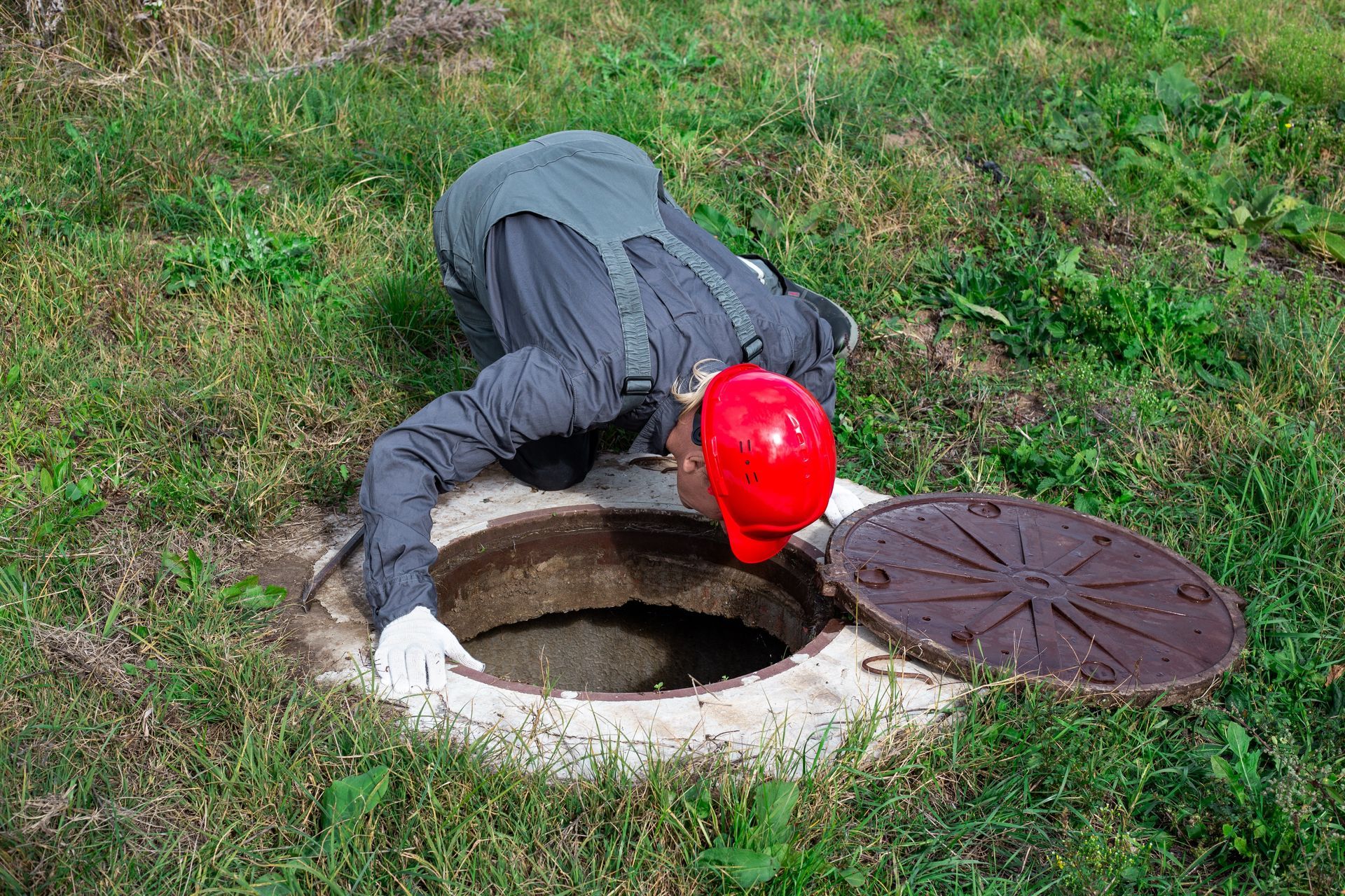 Worker in a red hard hat peering into an open manhole in a grassy area