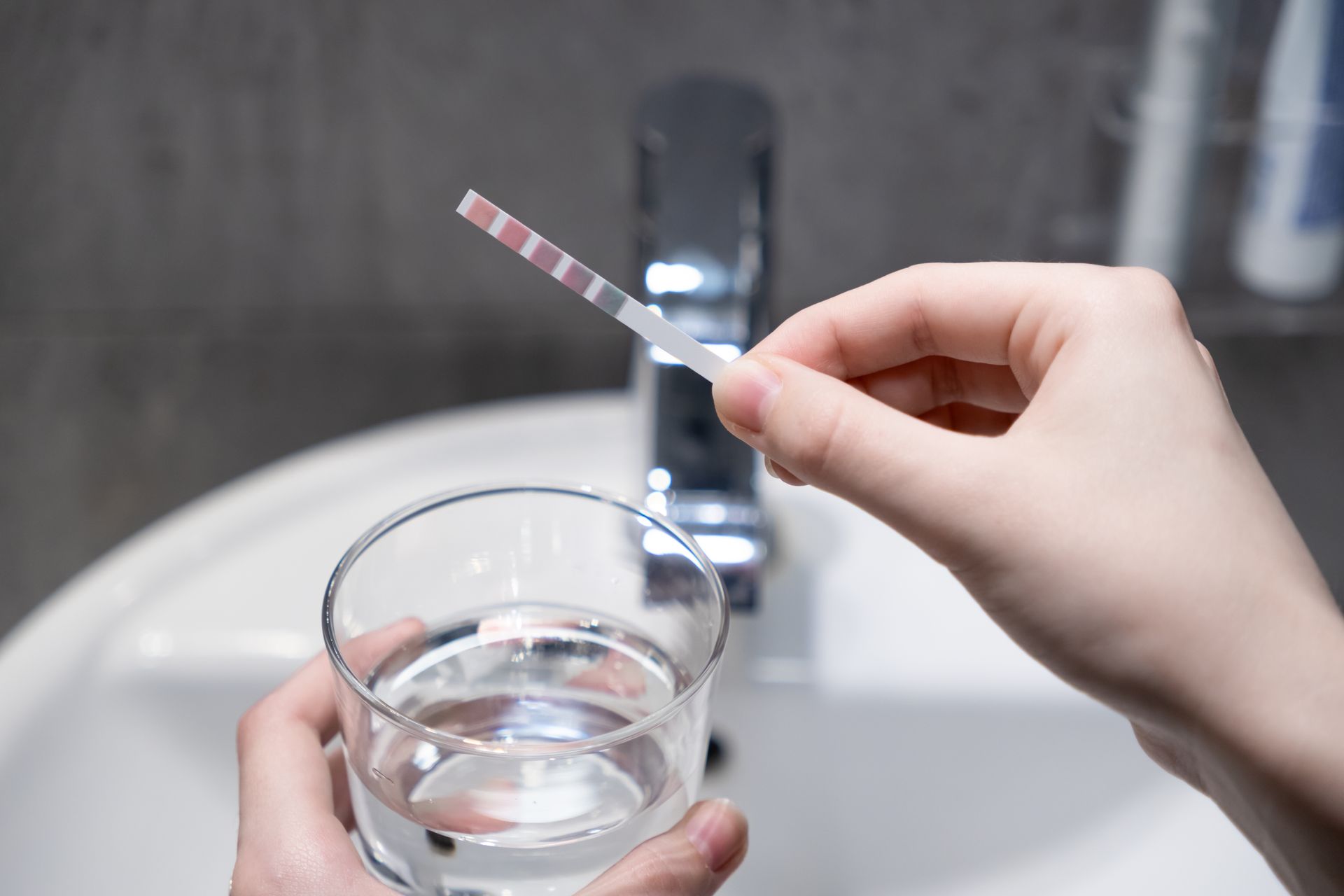 Hand holding a water glass while testing tap water with a strip at a bathroom sink