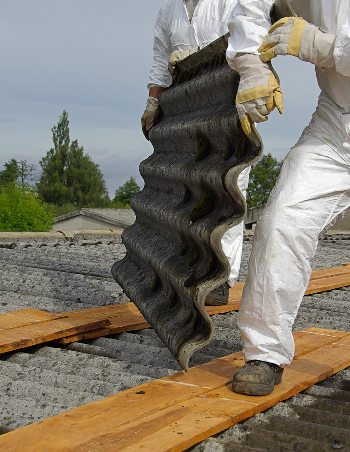 Worker in white protective suit handling a large black roofing shingle outdoors.