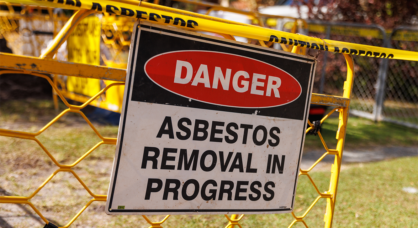 Warning sign reading “Danger: Asbestos Removal in Progress” beside yellow caution tape and fencing.
