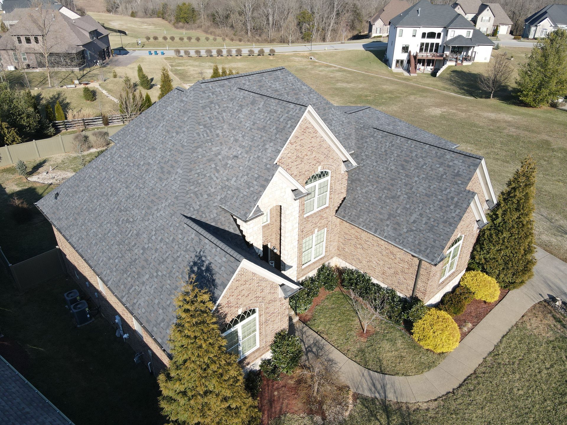 Aerial view of a suburban two-story brick house with a gray shingle roof, landscaped yard, and paved walkway.
