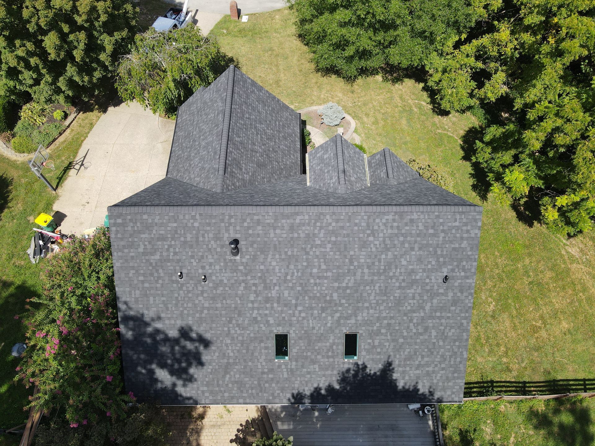 An aerial view of a dark shingled roof on a house, surrounded by trees and a green lawn.