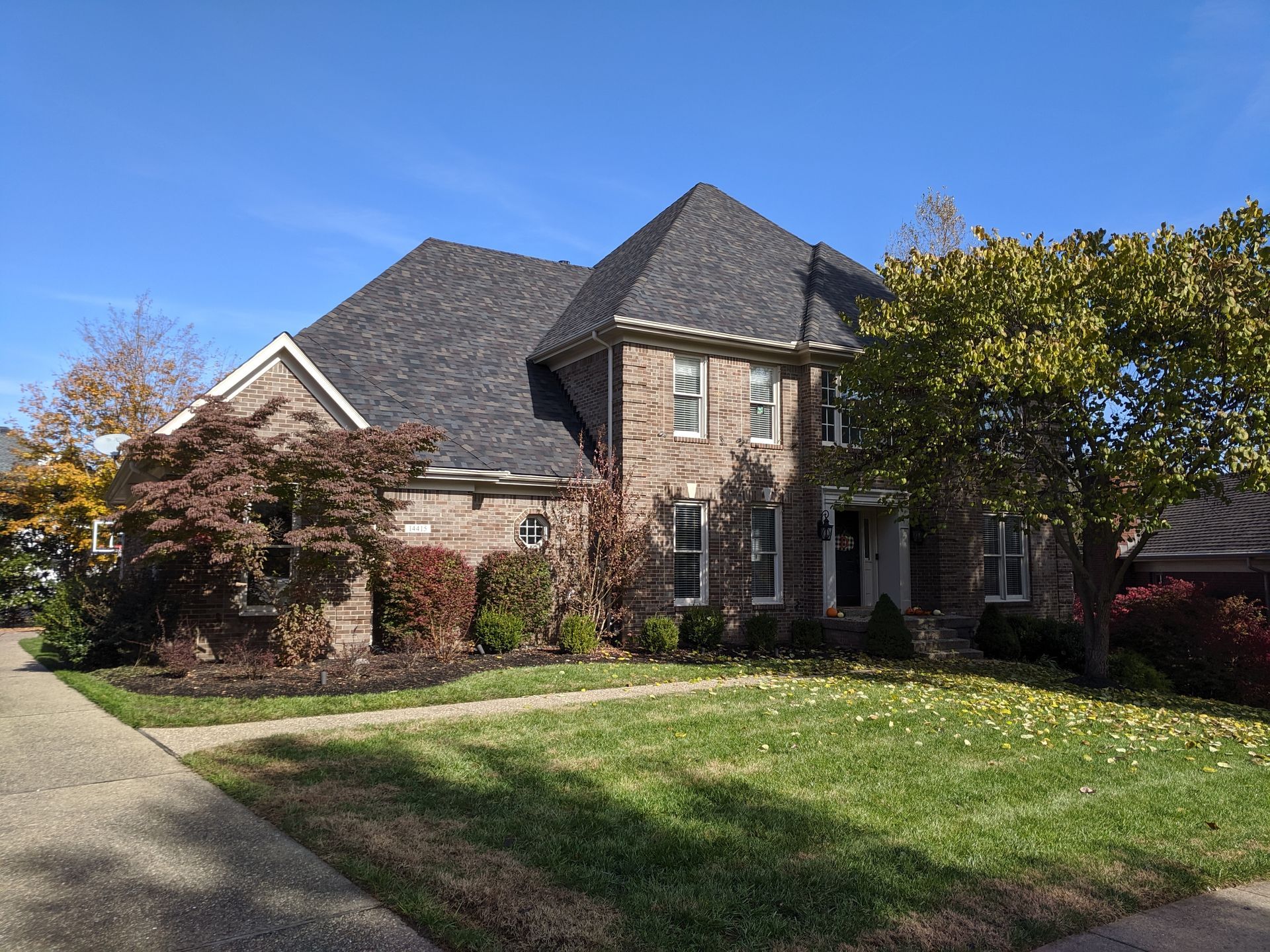 A two-story brick house with a dark gabled roof, surrounded by green lawns and trees under a clear blue sky.