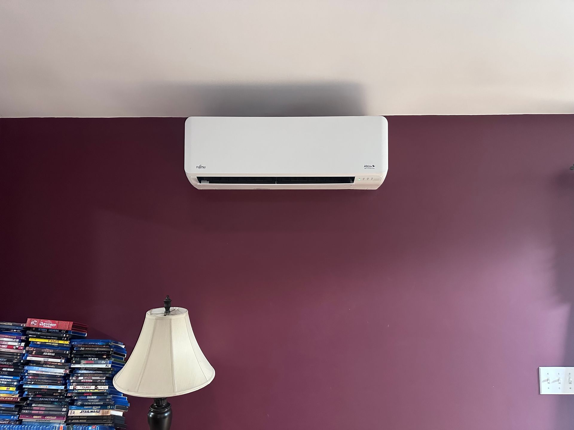 White wall-mounted air conditioner on a purple wall above a lamp and stacked items