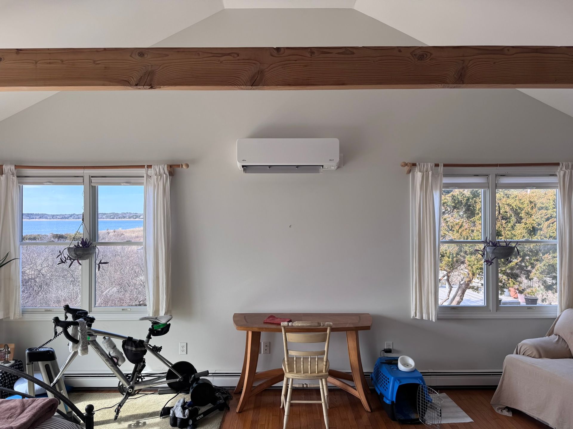 Bright living room with two windows, wall AC, wooden beam, and a bicycle near a small table and couch.