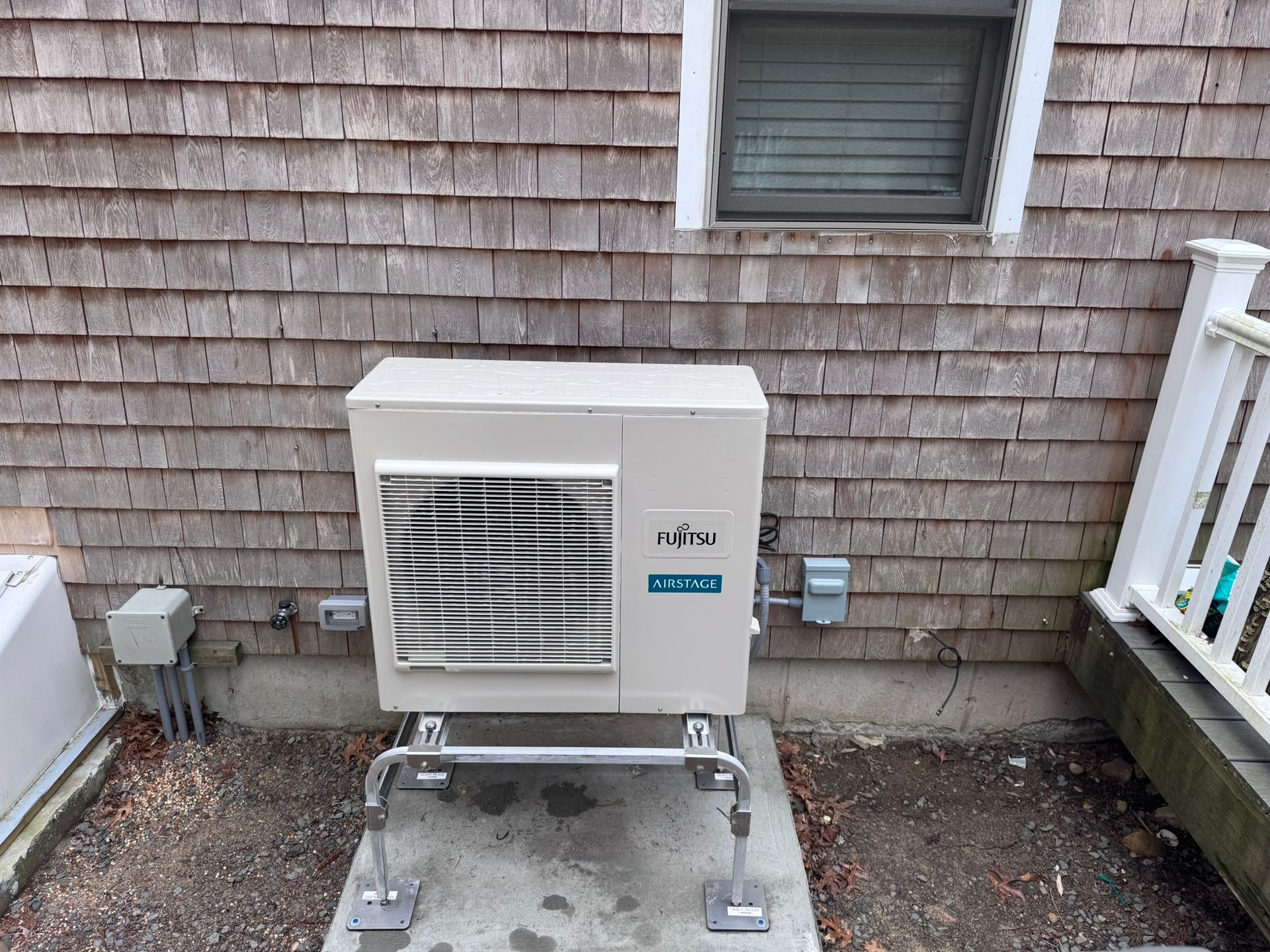 A white air conditioner is sitting outside of a house next to a window.