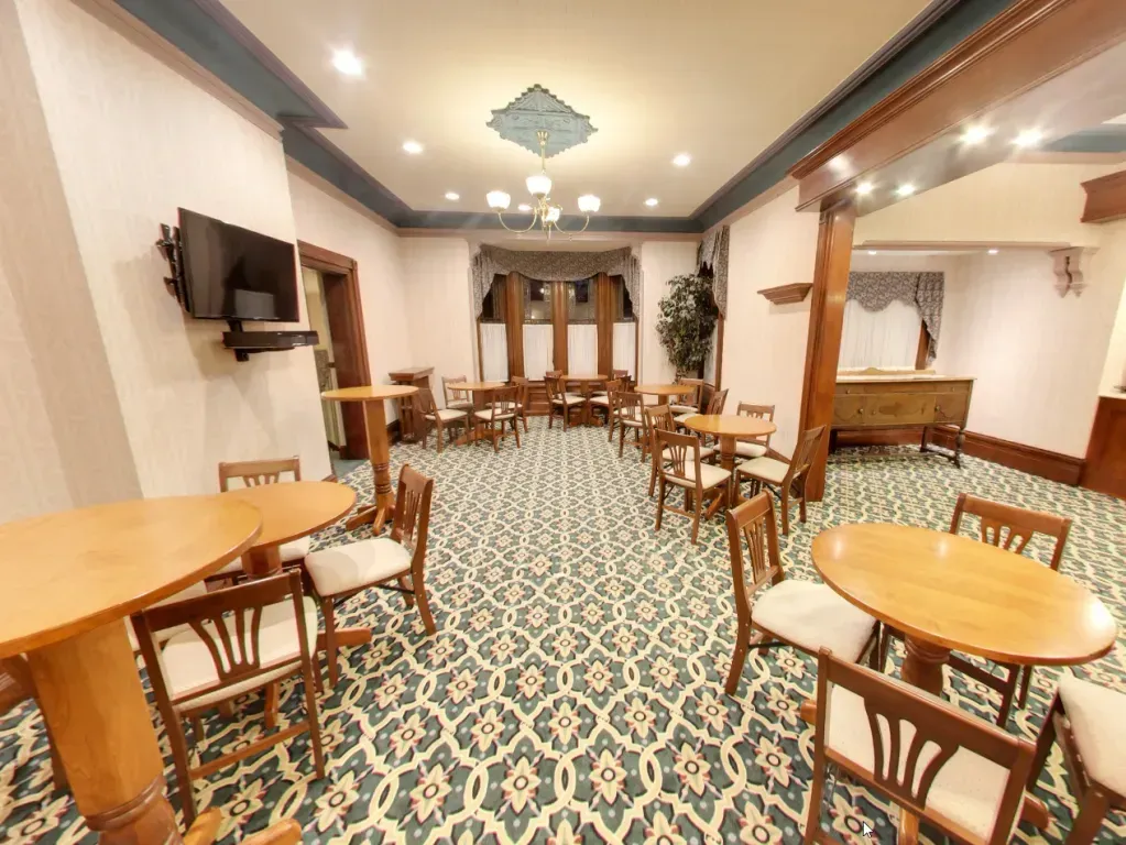 Empty restaurant dining room with round wooden tables, chairs, patterned carpet, and ceiling lights