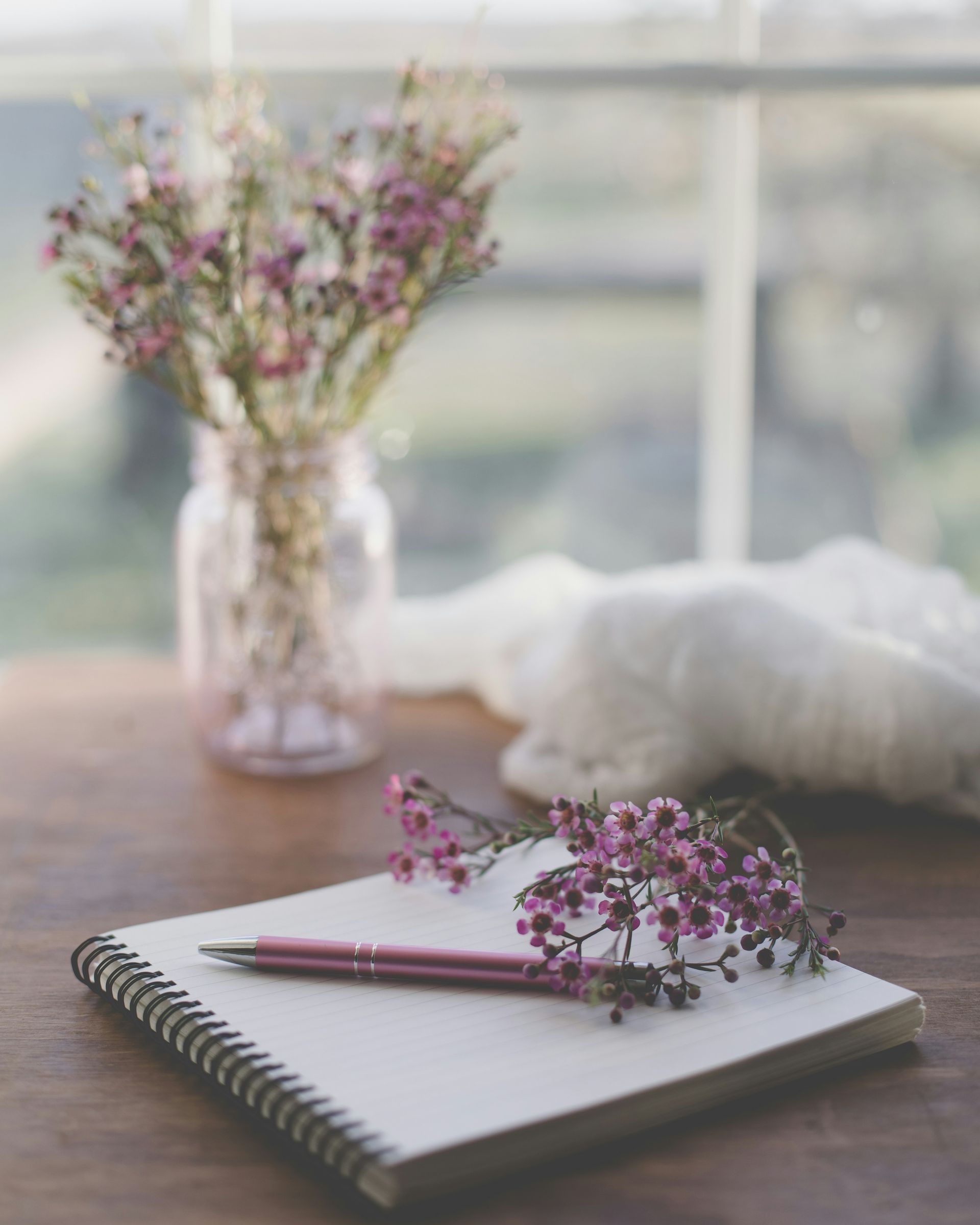Notebook with pink pen and flowers on a wooden table by a bright window