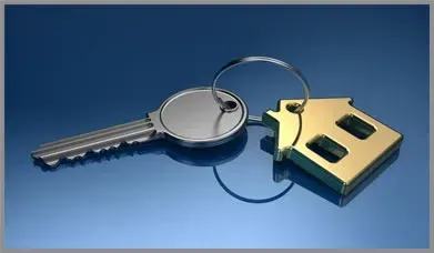 Silver key on a keyring next to a brass house-shaped keychain on a blue background