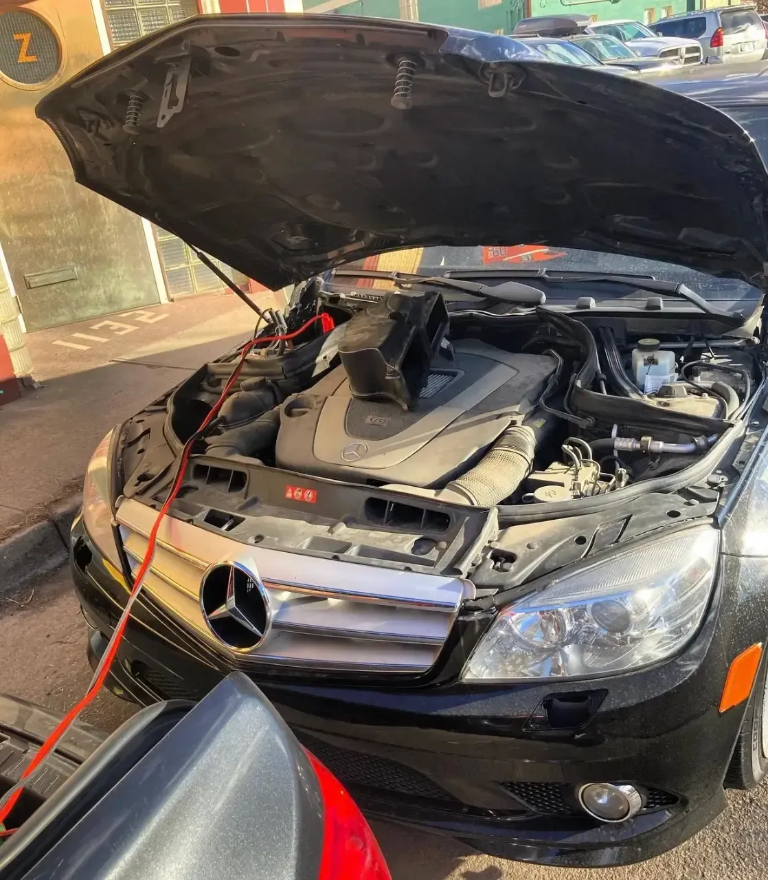 Black Mercedes sedan with hood open, jumper cables attached in a garage or service area