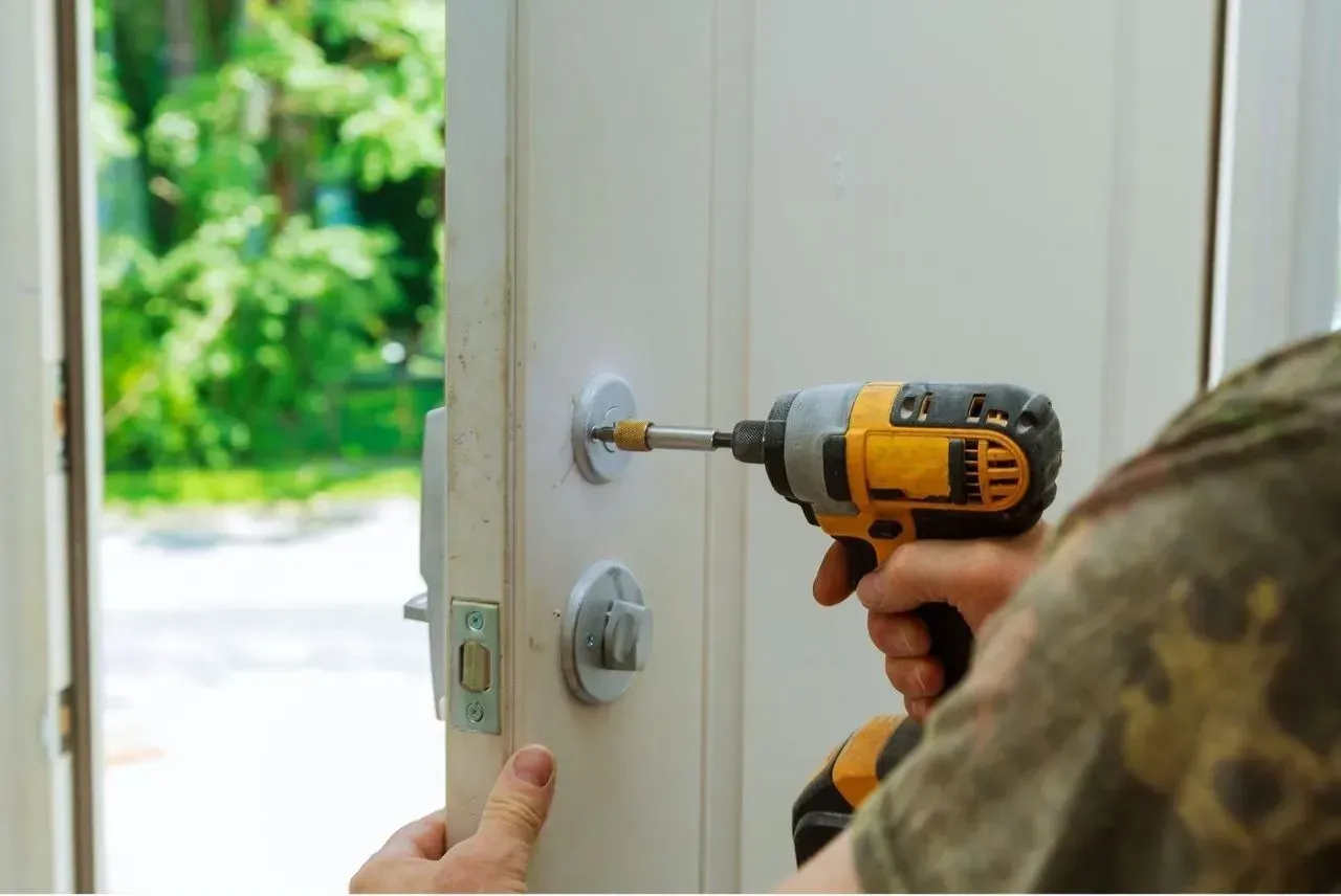 Person using a cordless drill to install a lock on a white door, with greenery outside.