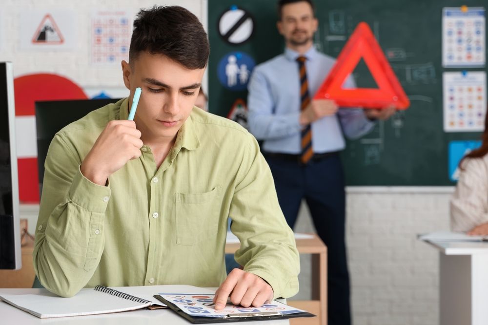 Student studying at a desk while a teacher stands in the classroom behind him, holding a red triangle sign.