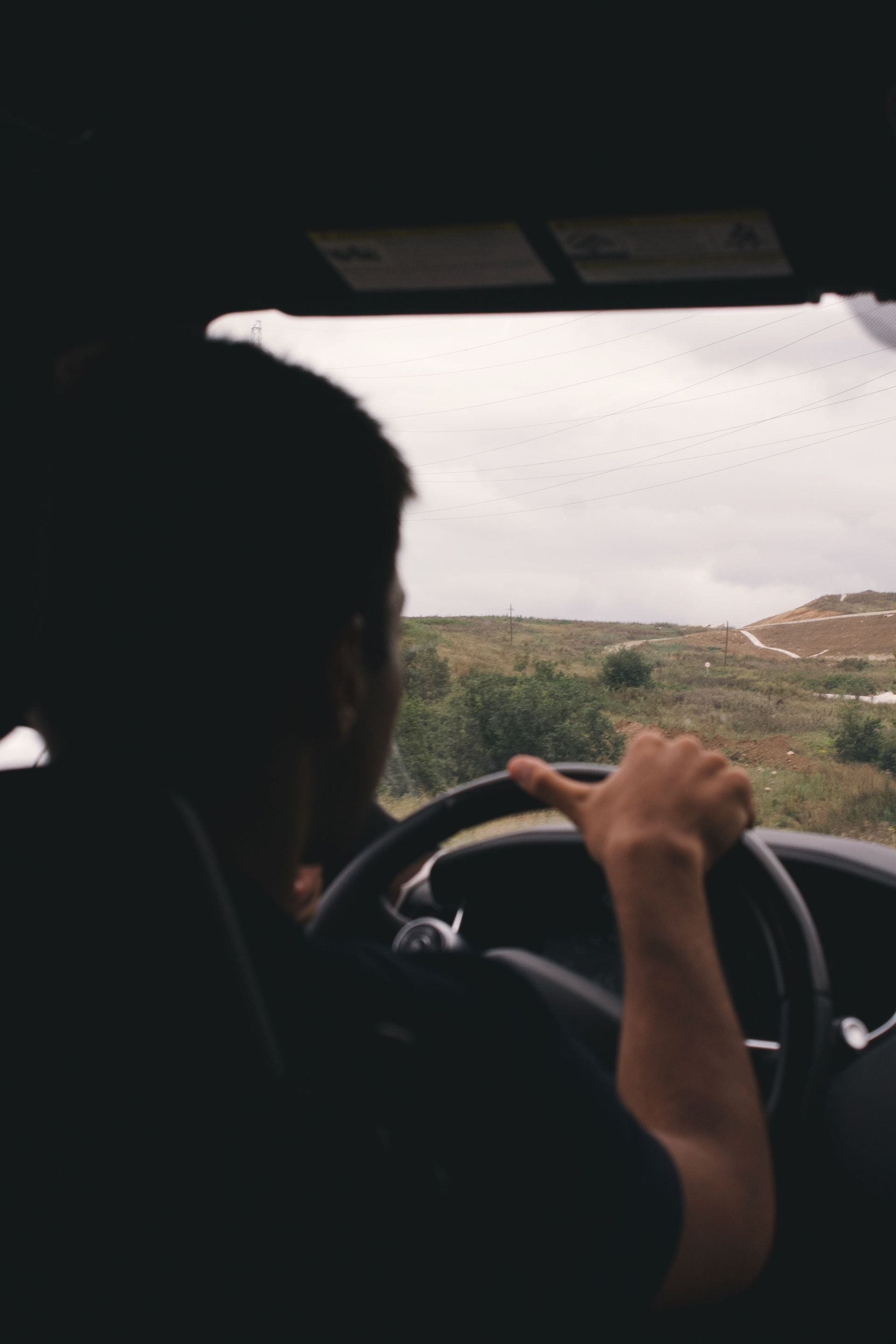 Driver seen from behind steering a car on a rural road under an overcast sky