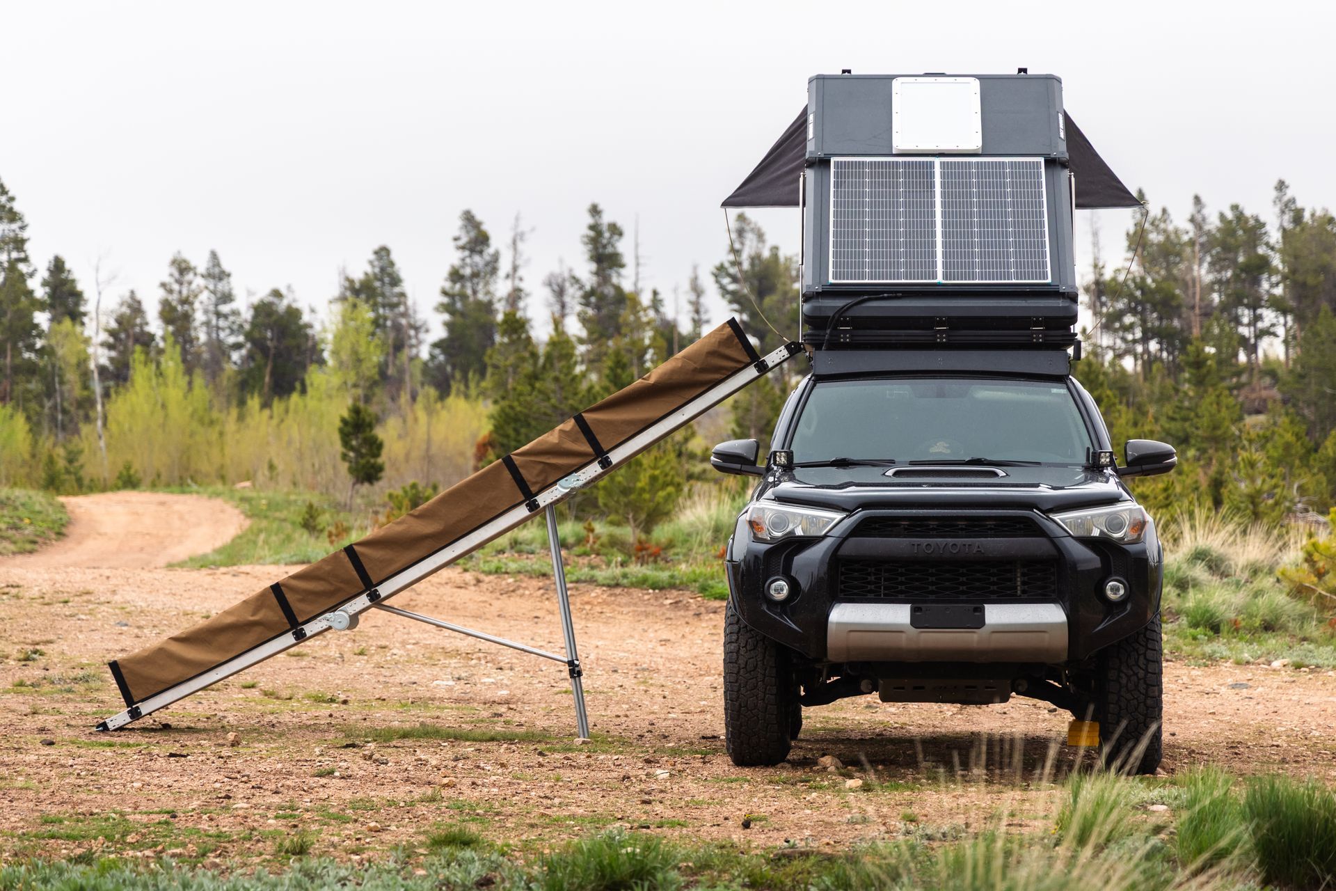 Black SUV with rooftop tent and awning parked on a dirt road in a forest.