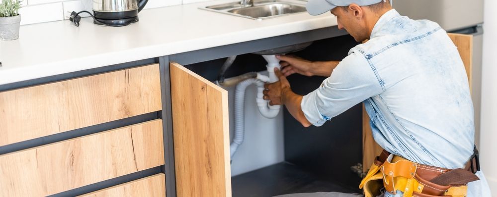 A plumber in a light blue shirt works under a kitchen sink. Wooden cabinet doors are open.