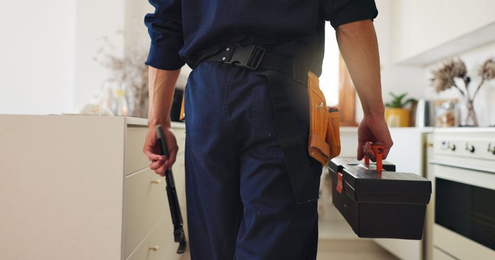 Plumber in navy blue uniform carrying toolbox and wrench in kitchen.