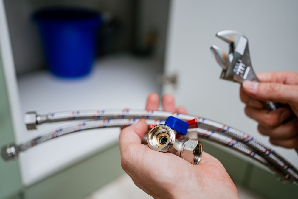 Hands holding plumbing parts, including a wrench, flexible tubes, and valves, in front of an open cabinet.