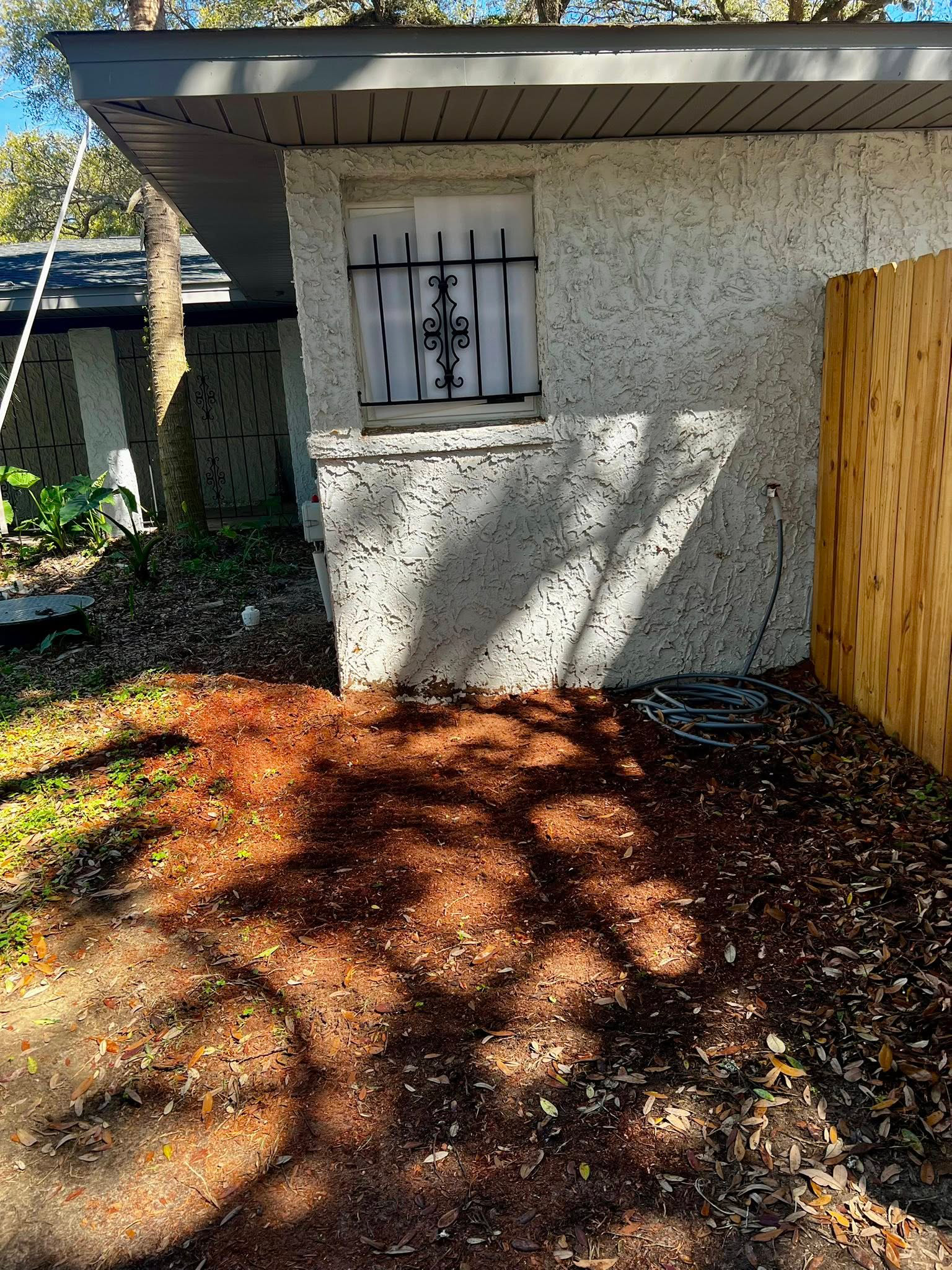 A light-colored stucco building with a window protected by a decorative metal grate, set next to a wooden fence.