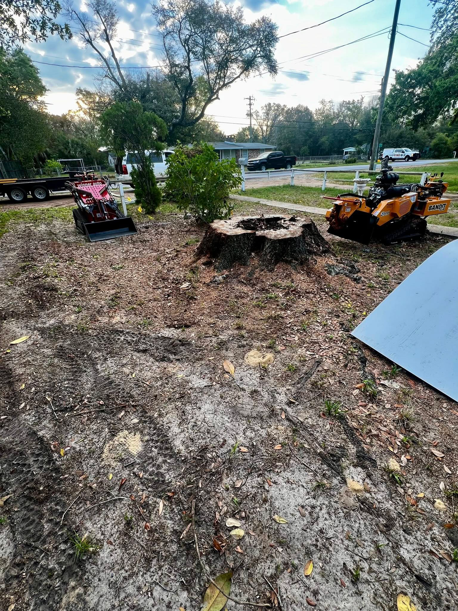 A pink and a yellow ATV sit on a leaf-covered lawn next to a tree stump, with houses and vehicles visible in the background.