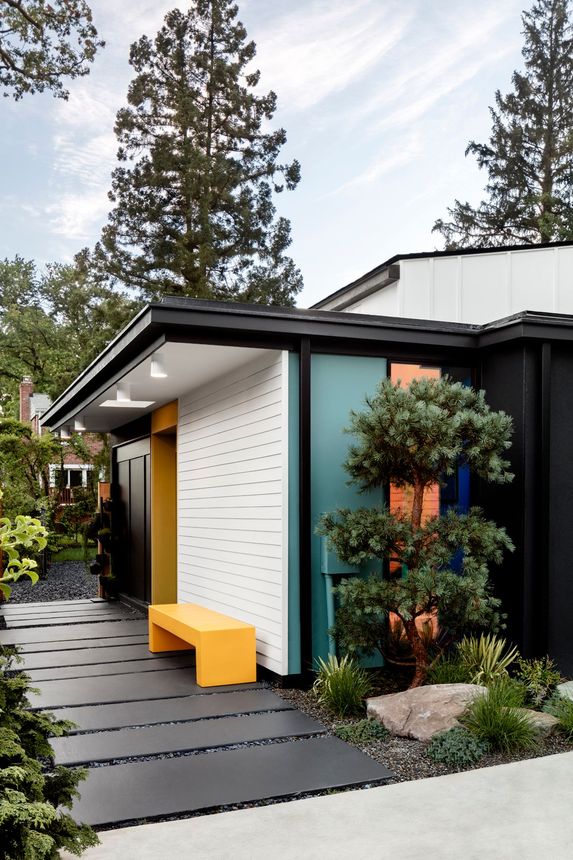 Modern house entrance with white brick, black trim, yellow bench, and potted trees along a stone path