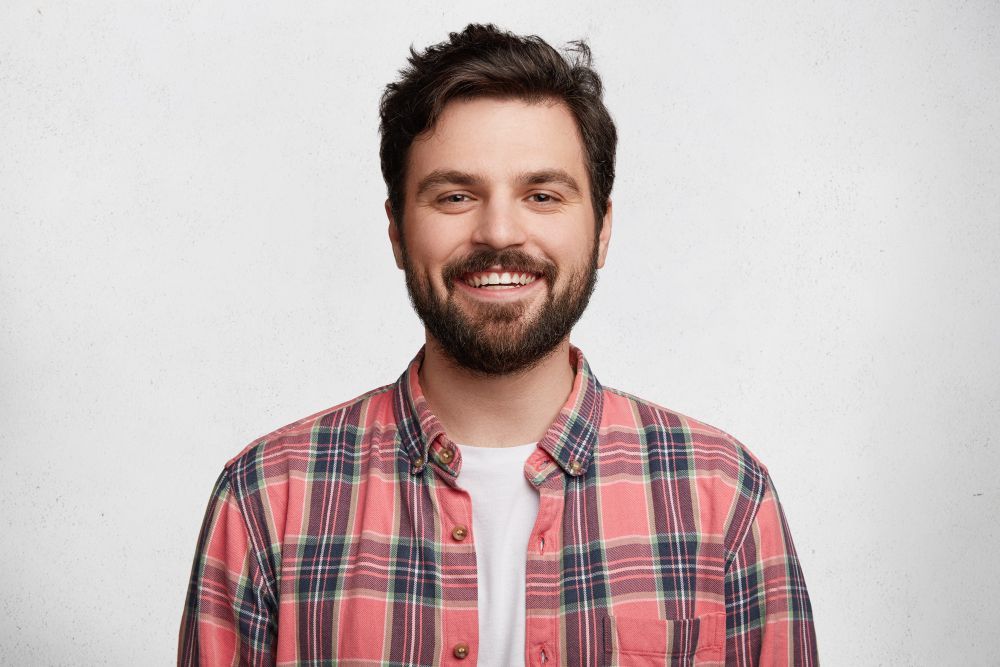 Smiling bearded man in a red plaid shirt against a light gray background