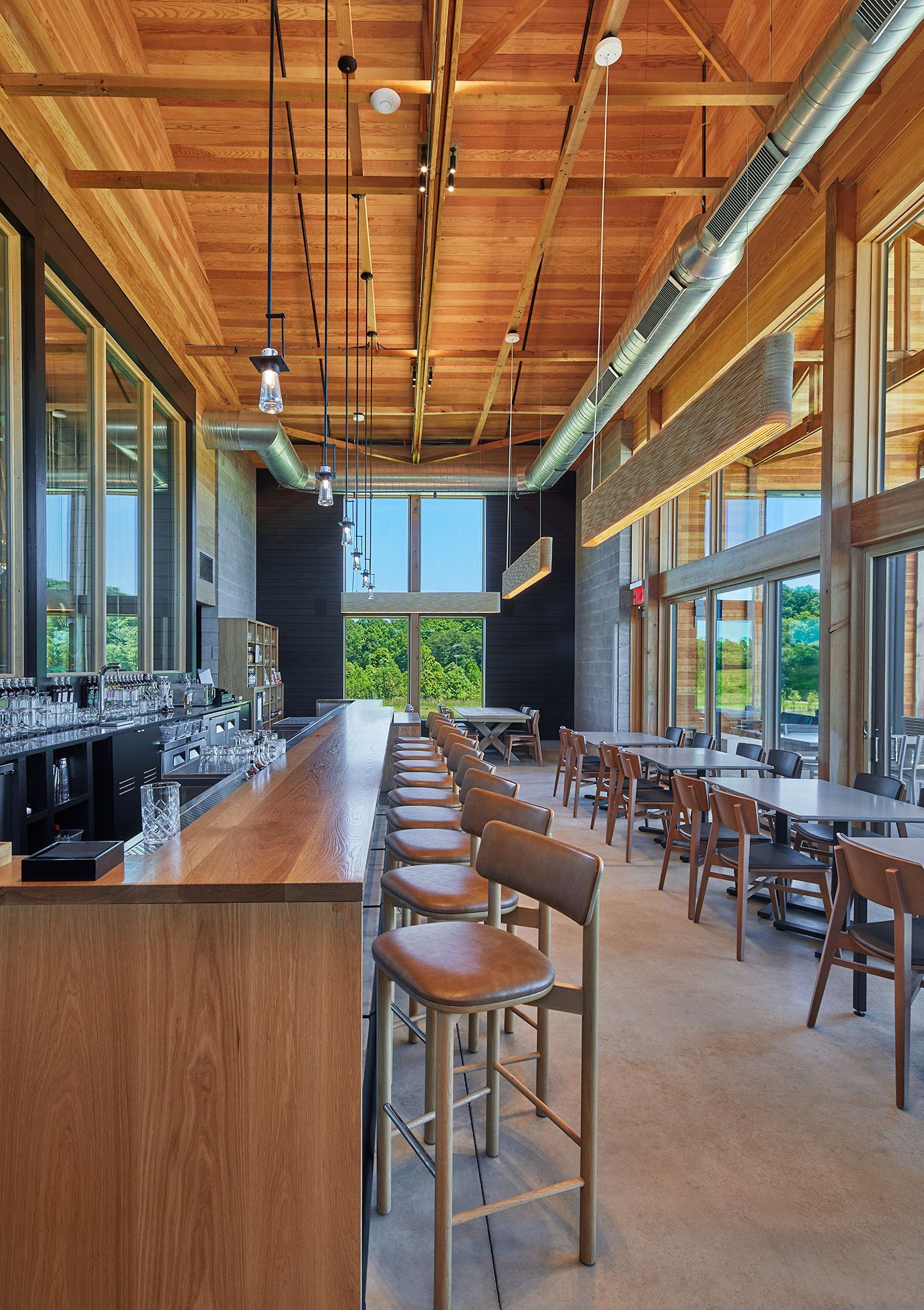 Modern restaurant interior with wooden bar, high stools, and large windows under a vaulted wood ceiling