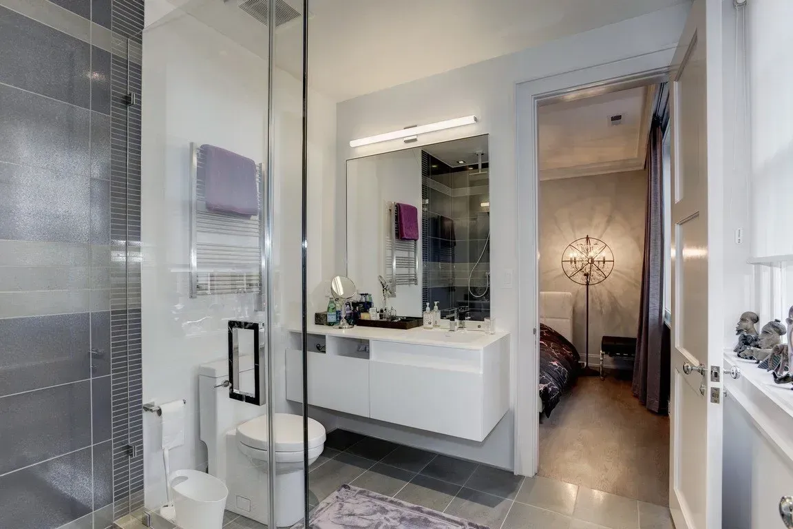 Modern bathroom with glass shower, toilet, vanity sink, and doorway to a hallway with a chandelier.