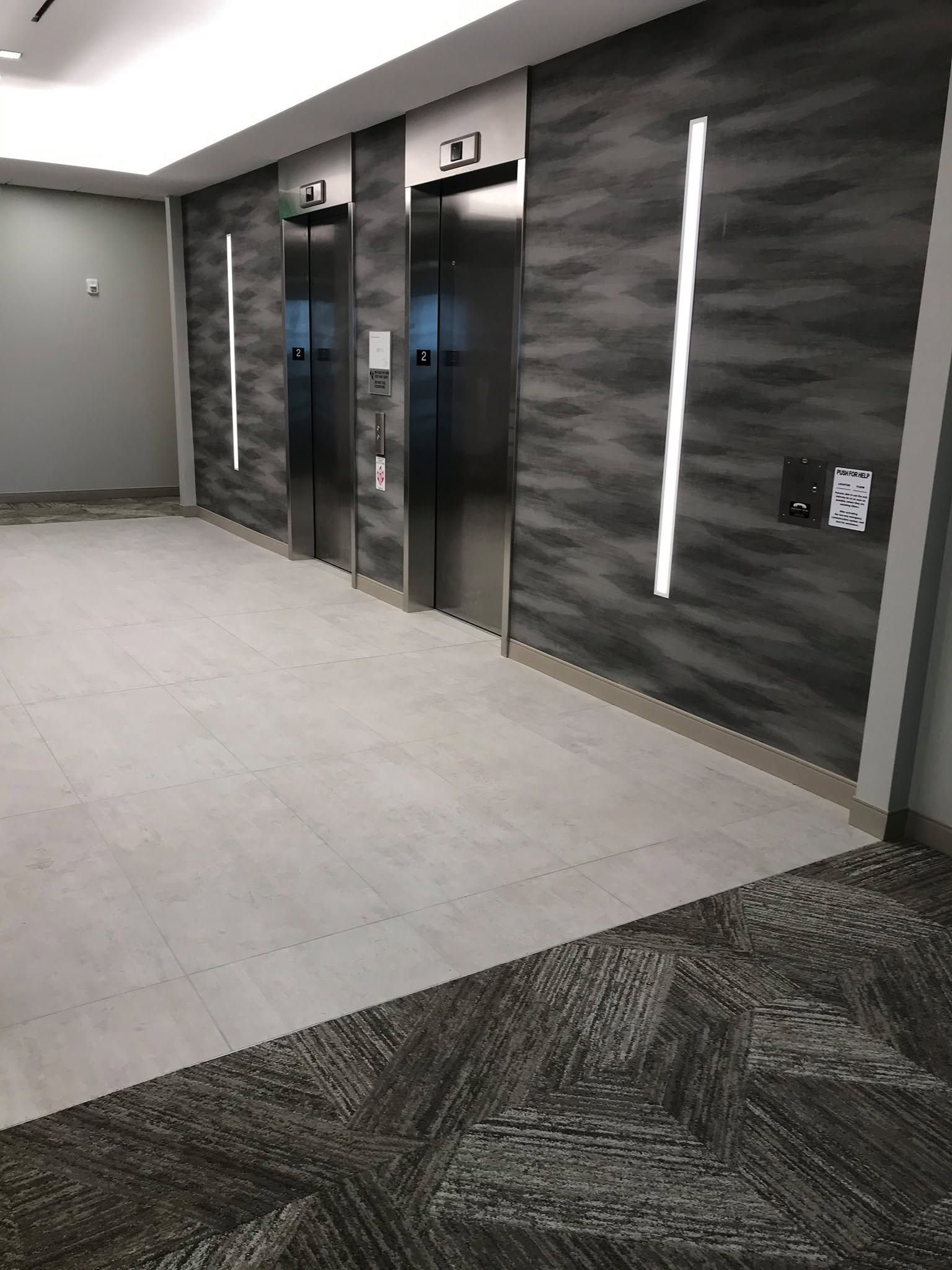 Modern hallway with elevator doors, gray tiled floor, and dark stone-pattern wall panels.