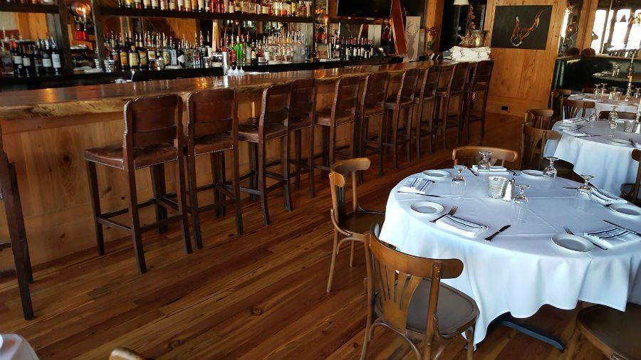 Empty restaurant dining room with wooden bar stools, tables set with white tablecloths, and a polished wood floor
