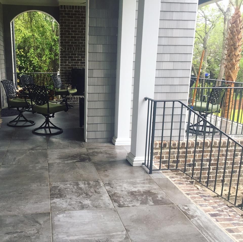 Covered porch with gray tile floor, black patio railing, and brick archway overlooking a wooded yard