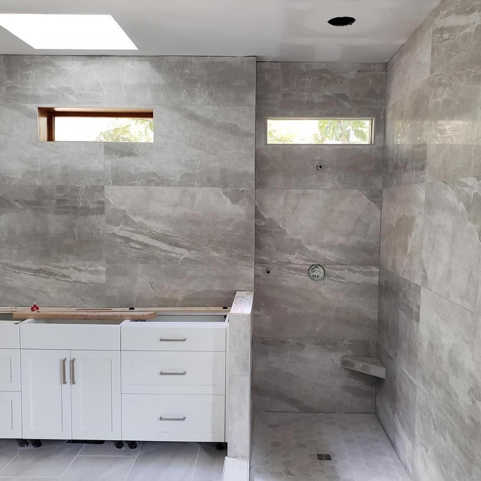 Unfinished bathroom with gray tile walls, walk-in shower, and white vanity under two small windows.