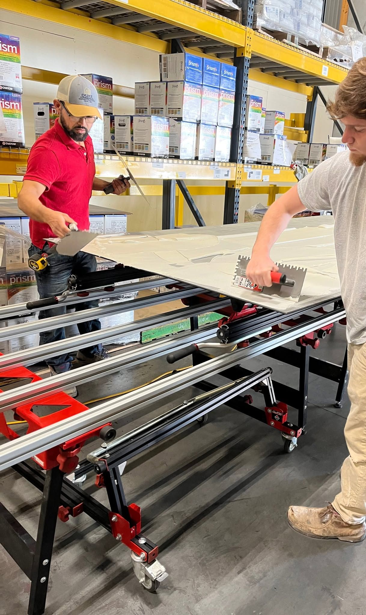 Two workers handling a large sheet of material on a metal assembly table in a warehouse.