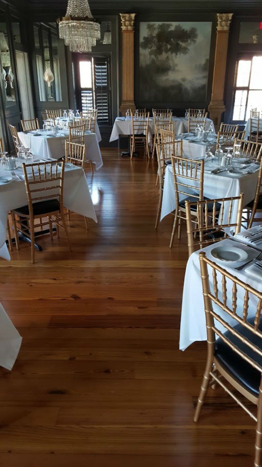 Empty formal dining room with round tables, gold chairs, white linens, and a polished wooden floor.